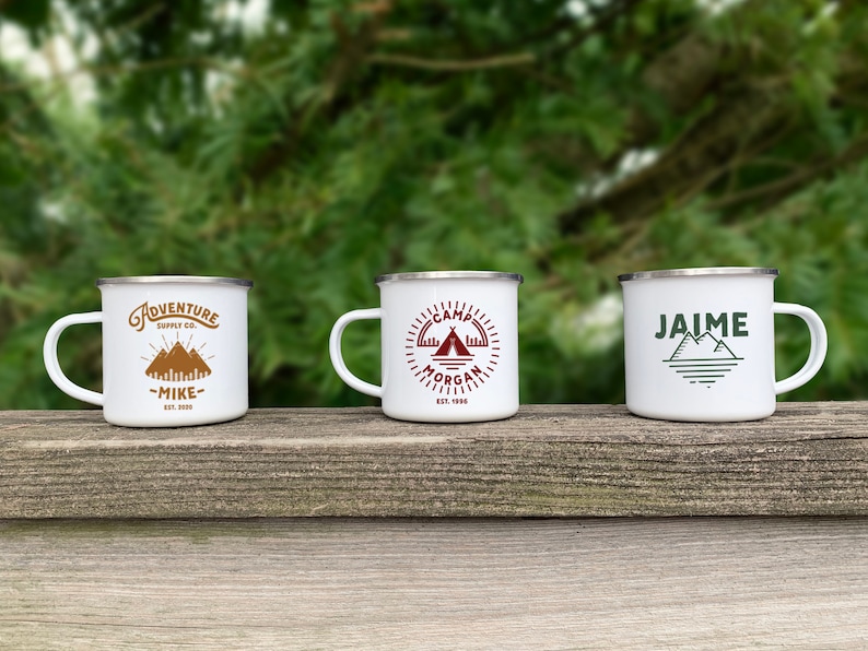 K&ouml;nnte beinhalten: Drei wei&szlig;e Emaille-Campingbecher mit verschiedenen Designs. Der erste Becher hat ein braunes Bergdesign mit dem Text "Adventure Supply Co. -Mike- Est. 2000". Der zweite Becher hat ein rotes Zeltdesign mit dem Text "Camp Morgan Est. 1990". Der dritte Becher hat ein gr&uuml;nes Bergdesign mit dem Text "Jaime".