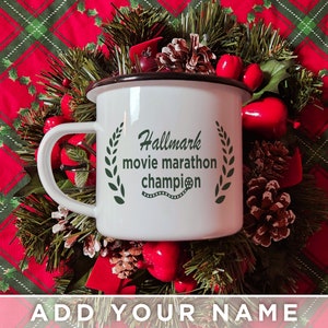 May include: White enamel mug with a black rim and handle. The mug has a green wreath design with the text "Hallmark movie marathon champion" and a film reel symbol.