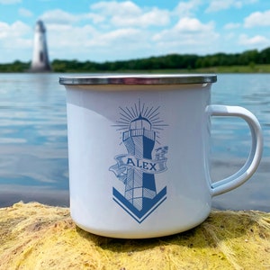 May include: White enamel camping mug with a blue lighthouse design and the name "ALEX" on a banner. The mug has a silver rim and handle.