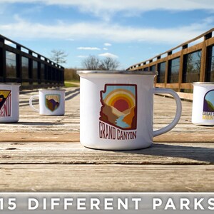 May include: Four white enamel mugs with colorful designs of national parks. The mugs feature the text "Big Bend", "Grand Canyon", "Great Smoky Mountains", and a silhouette of a mountain range. The mugs are arranged on a wooden surface.