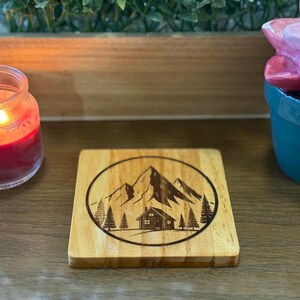 May include: Square wooden coaster with a mountain and cabin design etched in a circular frame. The coaster is light brown with a natural wood grain. A lit red candle and a succulent plant are in the background.