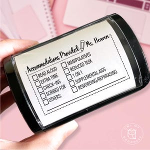 May include: A black stamp with a clear window displaying a checklist titled "Accommodations Provided." The checklist includes options like "Read Aloud," "Extra Time," and "Manipulatives." The stamp is held against a pink background.