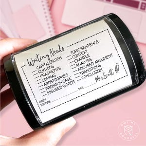 May include: A black rectangular stamp with a white label featuring the words "Writing Needs" and a list of writing topics. The stamp includes a space for a parent's signature and the date. The stamp is held in a person's hand.