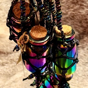 May include: Three iridescent glass bottles with cork stoppers are suspended by black netting and beaded strands. The bottles reflect a spectrum of colors, including purple, green, and blue. The background is a soft, neutral tone.