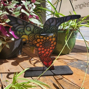 Op de afbeelding: Een kleurrijke houten kolibrie met een mandala-ontwerp, staand op een zwarte houten basis. De kolibrie is geschilderd in tinten blauw, groen, geel, oranje en rood.