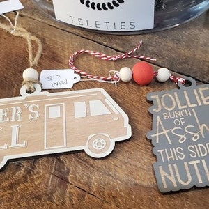 Op de afbeelding: Twee houten kerstversieringen met humoristische spreuken. De eerste versiering is vanvormig en zegt "Shitter's Full". De tweede versiering is grijs en zegt "Jolliest Bunch of Assholes This Side of the Nuthouse".