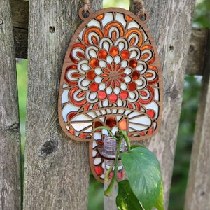 Op de afbeelding: Een houten wandhanger in de vorm van een paddenstoel met een rood en wit mandala-ontwerp. Een doorzichtige glazen buis met een plant hangt aan de onderkant van de paddenstoel.