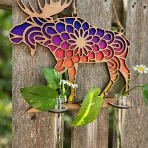 Op de afbeelding: Houten eland wanddecoratie met een mandala-ontwerp in tinten paars, oranje en geel. De eland heeft drie heldere glazen reageerbuizen om bloemen of groen in te houden.