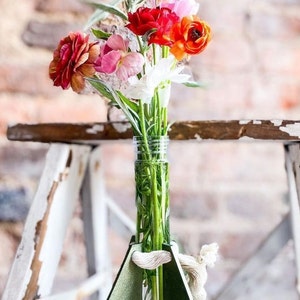 Op de afbeelding: Een levendig bloemstuk in een heldere glazen vaas, met een verscheidenheid aan kleurrijke bloemen. De vaas wordt vastgehouden door een groene geometrische houder met een touwaccent. De bloemen omvatten roze, rode en oranje bloesems.