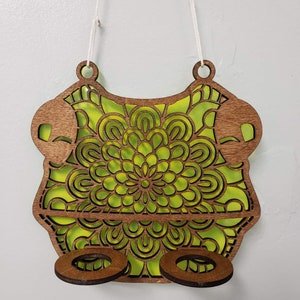 Op de afbeelding: Een houten wandhanger met een groen mandala-ontwerp en twee ronde uitsparingen. De hanger is bevestigd aan een wit touw.