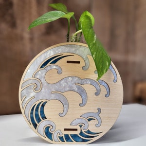 Op de afbeelding: Een houten vaas met een golfontwerp in blauw en wit. De vaas is gemaakt van licht hout en heeft een ronde vorm. Een kleine plant staat in de vaas.