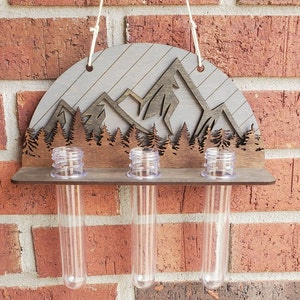 Op de afbeelding: Houten wanddecoratie met een berg- en bosontwerp. Het heeft drie heldere glazen reageerbuizen om kleine planten of bloemen in te bewaren.
