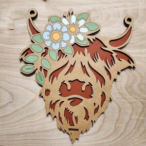 May include: A wooden cutout of a Highland cow's head with a floral crown. The cow is brown with a light brown muzzle and white eyes. The flowers are white, light blue, and yellow with green leaves.