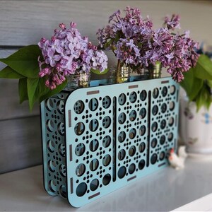 Op de afbeelding: Een lichtblauwe houten bloempot met een geometrisch patroon. De pot is gevuld met paarse lila bloemen.