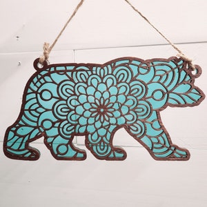 May include: A wooden bear silhouette with a teal mandala design. The bear is standing on its four legs and has a detailed design.