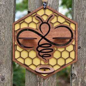 Op de afbeelding: Houten wanddecoratie in de vorm van een bij met een honingraatpatroon en een kleine ring aan de onderkant. De bij is bruin met zwarte details en is zwart omrand. De honingraat is geel.