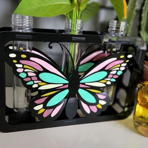 Op de afbeelding: Zwarte houten vlinder-vormige bloempot met drie heldere glazen reageerbuizen om bloemen in te houden. De vaas staat op een wit oppervlak.