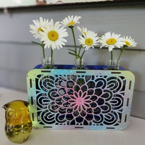 Op de afbeelding: Een houten bloemenvaas met een blauw, roze en groen mandala-ontwerp. De vaas heeft vier kleine glazen buisjes om bloemen in te houden. Witte madeliefjes staan in de vaas. Een geel glazen uilbeeldje staat op de voorgrond.