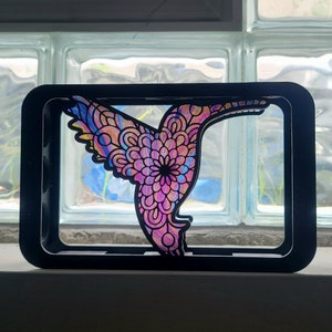 Op de afbeelding: Een zwart frame met een kleurrijke kolibrie-uitsparing. De kolibrie is roze, paars, blauw en geel met een mandala-achtig ontwerp.