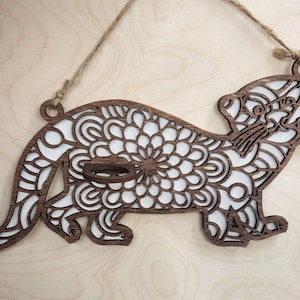 Op de afbeelding: Een houten uitsnijding van een otter met een mandala-ontwerp. De otter is bruin met witte details en heeft een gat bovenaan om op te hangen.