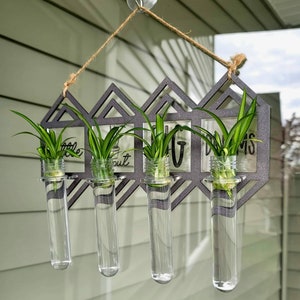 Op de afbeelding: Een hangende plantenhanger met vier glazen reageerbuizen gevuld met kleine planten. De plantenhanger is gemaakt van hout en heeft een geometrisch ontwerp. De tekst "Little Dreams But Big Dreams" is op de plantenhanger gedrukt.