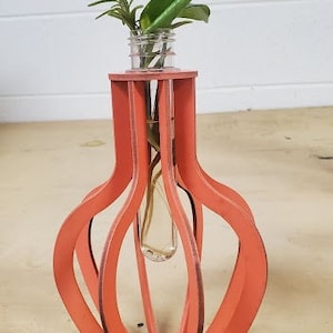 Op de afbeelding: Een koraalkleurige houten vaas met een geometrisch ontwerp bevat een glazen reageerbuis met een kleine plant erin.