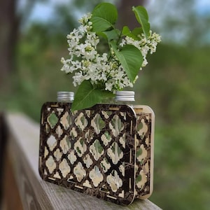 Op de afbeelding: Een houten bloemenvaas met een geometrisch patroon in bruin en zwart. De vaas heeft drie glazen cilinders om bloemen in te houden. Een boeket witte lila bloemen staat in de vaas.