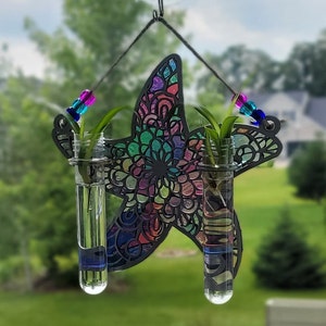 Op de afbeelding: Een kleurrijke, mandala-stijl plantenhanger in de vorm van een zeester met twee glazen reageerbuizen gevuld met water en kleine planten. De hanger is gemaakt van zwart metaal en heeft een kralenketting om op te hangen.