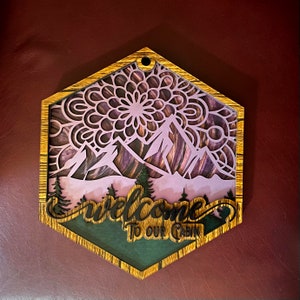 Op de afbeelding: Houten zeshoekig bord met een paars en zwart mandala-ontwerp, bergen en bomen. Het bord zegt "Welkom in onze hut" in zwarte letters.