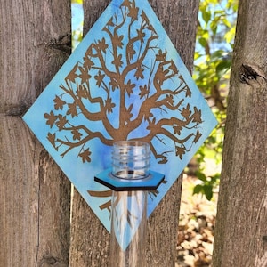 Op de afbeelding: Een houten, ruitvormige wandhanger met een blauwe aquarel achtergrond en een bruin silhouet van een boom met bladeren. Een heldere glazen buis is bevestigd aan de onderkant van de hanger.