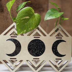 Op de afbeelding: Een houten plantenbak met drie halvemaanvormige openingen, elk gevuld met een zwart glitterachtig materiaal. De plantenbak heeft een geometrisch ontwerp en bevat drie kleine potplanten.