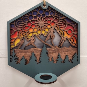 Op de afbeelding: Een teal-kleurige, zeshoekige houten wandhanger met een mandala-ontwerp in tinten blauw, rood, oranje en geel. De mandala bevindt zich achter een uitsnijding van een bergketen met een bos bomen aan de onderkant. De hanger heeft een teal-kleurige ring aan de onderkant om op te hangen.