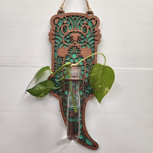 Op de afbeelding: Een teal en bruine houten wanddecoratie met een uitgesneden ontwerp van een otter. Een heldere glazen reageerbuisvaas is in de decoratie gestoken en bevat een kleine groene plant.