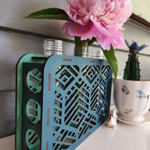 Op de afbeelding: Tealblauwe houten vaas met een geometrisch ontwerp en drie glazen cilinders om bloemen in te houden. Een roze pioenroos staat in de vaas.