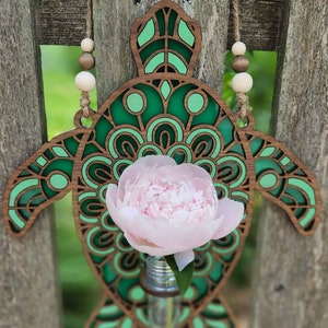 Op de afbeelding: Een houten wanddecoratie in de vorm van een schildpad met een groen mandala-ontwerp. Een roze pioenroos staat in een kleine glazen vaas in het midden van de schildpad.