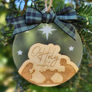 Op de afbeelding: Een houten kerstversiering met een matglazen achtergrond. De versiering toont een silhouet van de kerststal met de tekst "Oh Holy Night". De versiering is versierd met een groen-zwarte ruitjes strik.