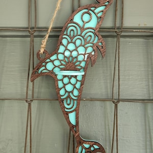 May include: A teal blue wooden dolphin with intricate cut-out designs. The dolphin is hanging from a twine string and has a small shelf in the middle.