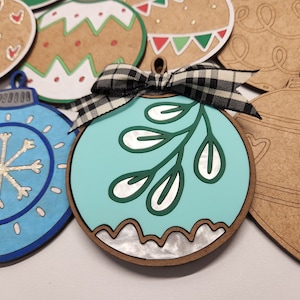 Op de afbeelding: Een set van zes onafgewerkte houten ornamenten, waaronder een blauwe ornament met een sneeuwvlokkenpatroon, een teal ornament met een groen bladontwerp en vier bruine ornamenten met verschillende ontwerpen.