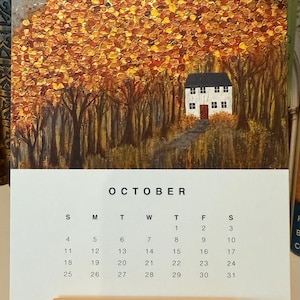 May include: A desk calendar for October featuring an autumn landscape painting with a white house and a path. The calendar is supported by a wooden stand. The word "OCTOBER" is printed at the top.