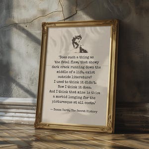 May include: A framed print with a gold ornate frame, featuring a quote from Donna Tartt's "The Secret History." The text is centered on a cream-colored background, with a small black and white image of a statue at the top.