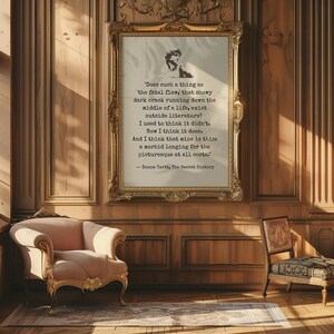 May include: A framed print with a gold ornate frame hangs on a wood-paneled wall. The print features a quote from Donna Tartt's "The Secret History" in black text. A pink armchair and a patterned chair sit on a rug.