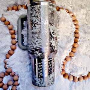 Puede incluir: Un vaso de acero inoxidable con un dise&ntilde;o detallado que presenta el rostro de una mujer, teclas de m&aacute;quina de escribir y recortes de peri&oacute;dicos. El vaso tiene un asa y una tapa. El texto "Polaris Belle Creative Co." es visible.
