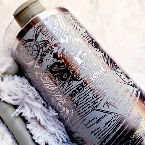 Puede incluir: Un vaso de acero inoxidable con pajita y tapa transparente. El vaso presenta un dise&ntilde;o detallado con texto e ilustraciones en plata y negro. El texto incluye las palabras "Torture Me Poet" y "Basement".