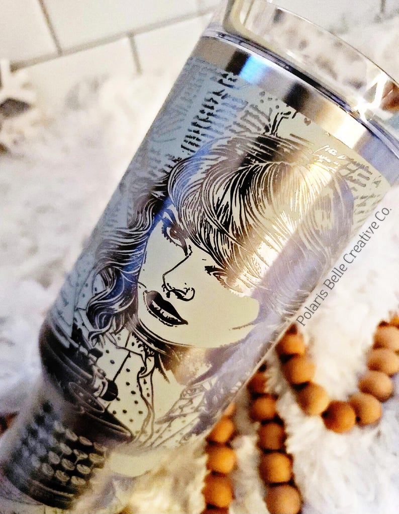Puede incluir: Un vaso plateado con un retrato grabado en blanco y negro de una mujer. El dise&ntilde;o se encuentra sobre un fondo de texto. Las palabras "Polaris Belle Creative Co." est&aacute;n impresas en el lateral del vaso. El vaso es cil&iacute;ndrico.