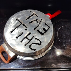 Puede incluir: Una tapa de metal plateado con un asa roja. La tapa tiene las palabras "THE FAT" recortadas.