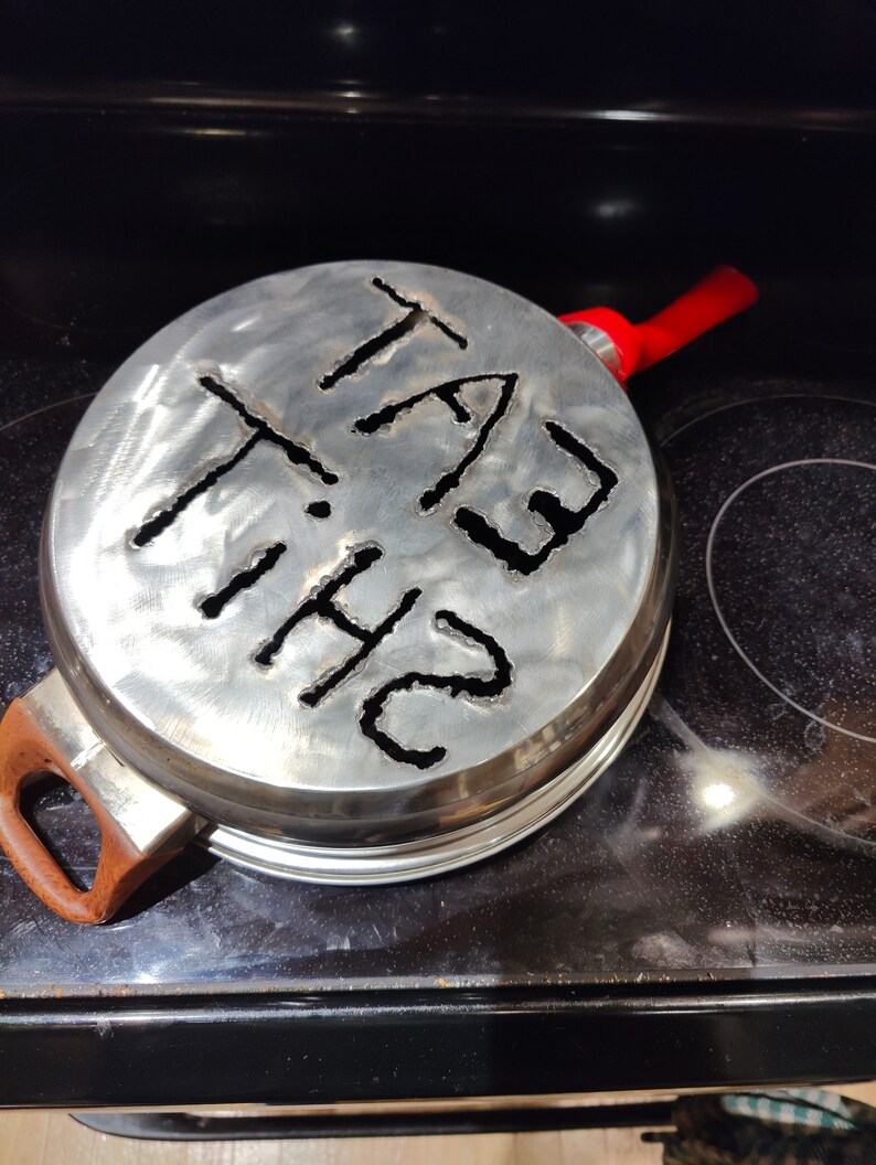 Puede incluir: Una tapa de metal plateado con un asa roja. La tapa tiene las palabras "THE FAT" recortadas.