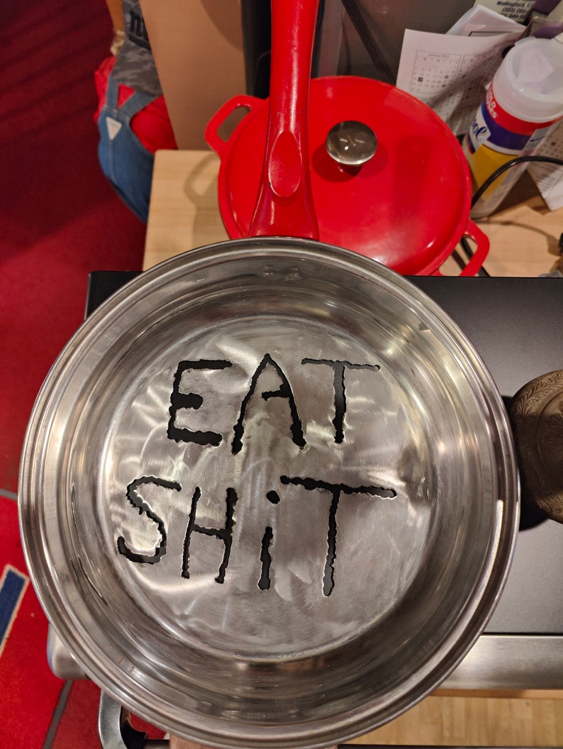 Puede incluir: Una sart&eacute;n de metal plateado con las palabras "EAT SHIT" grabadas en el fondo.