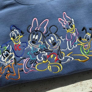 May include: A blue sweatshirt featuring embroidered images of Disney characters Mickey Mouse, Minnie Mouse, Donald Duck, Daisy Duck, and Pluto. The characters are outlined in various colors, including black, yellow, pink, and blue.