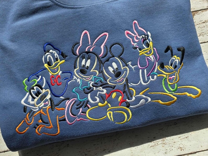 May include: A blue sweatshirt featuring embroidered images of Disney characters Mickey Mouse, Minnie Mouse, Donald Duck, Daisy Duck, and Pluto. The characters are outlined in various colors, including black, yellow, pink, and blue.