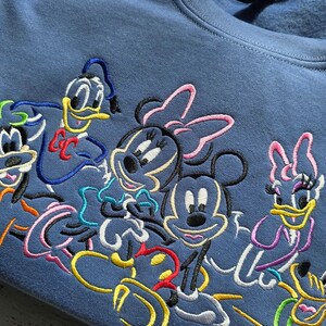 May include: A blue sweatshirt featuring embroidered cartoon characters including Mickey Mouse, Minnie Mouse, Donald Duck, Daisy Duck, and Goofy. The colorful embroidery is set against the solid blue fabric of the crew neck sweatshirt.